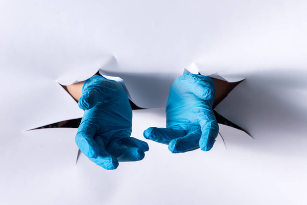 Mano femenina de un agujero en una pared de papel, con guantes médicos azules
 - Foto, imagen
