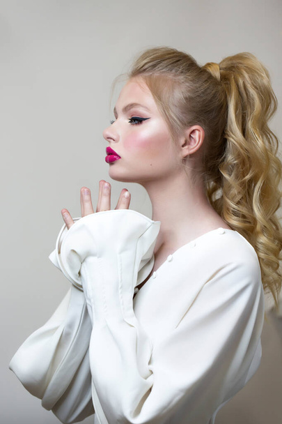 Portrait Woman in a white shirt with long magnificent sleeves. Bright makeup, blond hair gathered in a ponytail. Palms near the face. - Foto, imagen