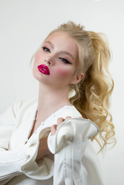 Woman model posing in studio on a white background. Clothing - a white blouse or shirt with a long sleeve. Blond hair is piled up in a ponytail. Professional bright makeup. Pink lips and black arrows. - Foto, Bild