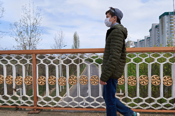 tiener jongen loopt door de straat gedurende de dag, een voetgangers loopbrug en hoogbouw met appartementen, een woonwijk, een medisch masker op zijn gezicht beschermt tegen virussen en stof - Foto, afbeelding