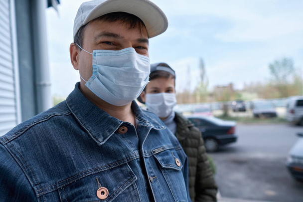 άνδρας και έφηβος αγόρι ποζάρουν κοντά σε τοίχο και κλειστή πόρτα πολυόροφων κτιρίων με διαμερίσματα, μια κατοικημένη περιοχή, μια ιατρική μάσκα στα πρόσωπά τους προστατεύει από ιούς και σκόνη - Φωτογραφία, εικόνα