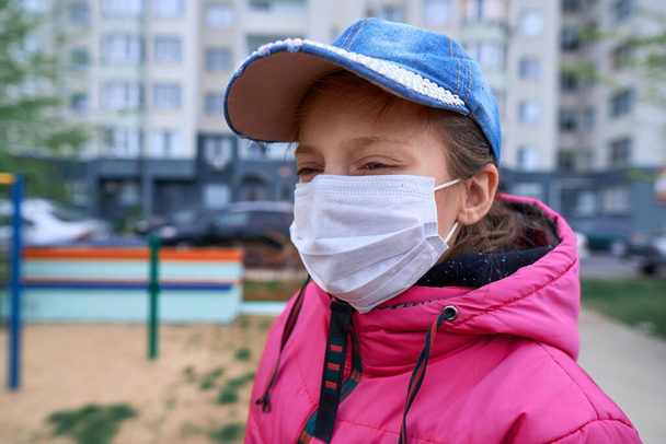 ένα κορίτσι που κάθεται σε μια τραμπάλα σε παιδική χαρά κοντά σε πολυώροφα κτίρια με διαμερίσματα, μια ιατρική μάσκα στο πρόσωπό της προστατεύει από ιούς και σκόνη - Φωτογραφία, εικόνα
