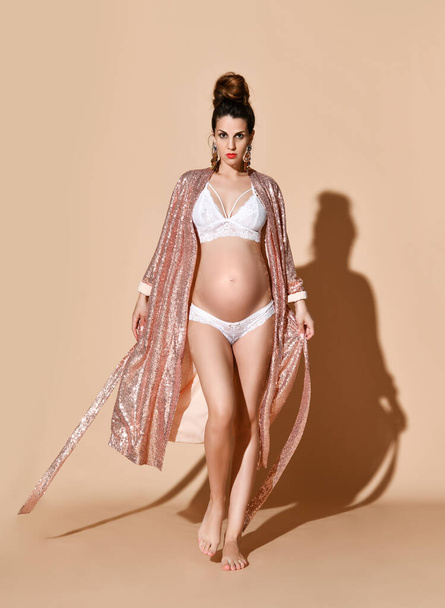 Pregnant female in earrings, white sexy lingerie and golden robe. She is walking against beige background. Full-length, copy space - Foto, Bild