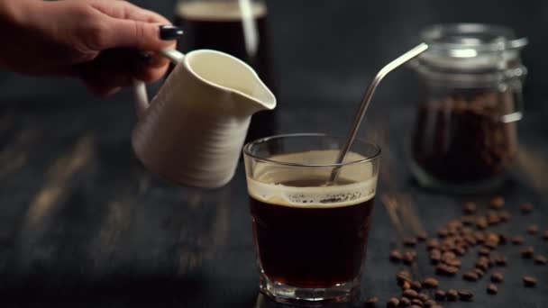 Slow motion of cream being poured into a glass of coffee on black wood table with metal cocktail tube and coffee beans on black background - Záběry, video