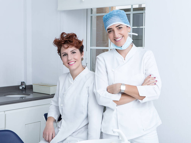 Ευτυχισμένες ενήλικες γυναίκες με ιατρική στολή χαμογελώντας και κοιτάζοντας την κάμερα ενώ εργάζονται στο σύγχρονο γραφείο στην οδοντιατρική κλινική - Φωτογραφία, εικόνα