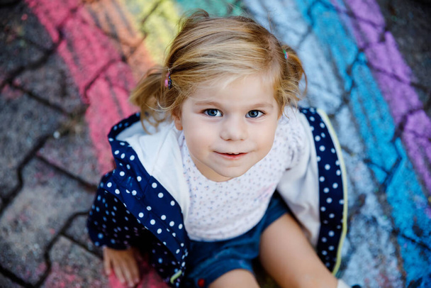 Szczęśliwa mała dziewczynka w gumowych butach z tęczowymi słońcem i chmurami z deszczem pomalowanymi kolorowymi kredkami na ziemi lub asfalcie w lecie. Cute dziecko zabawy. kreatywny wypoczynek - Zdjęcie, obraz