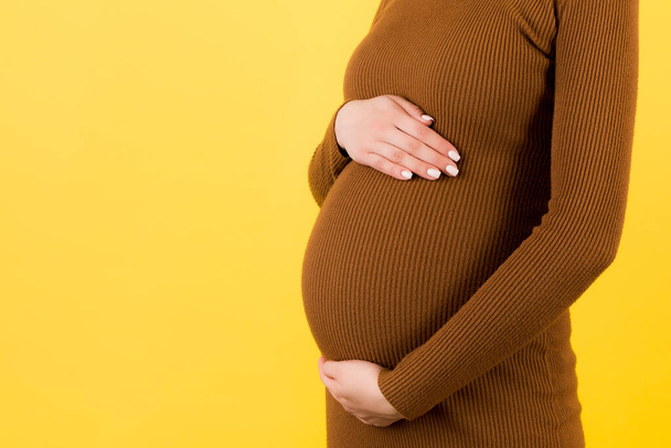 Обрезанное изображение беременной женщины в коричневом платье, прикасающейся к животу на желтом фоне. Будущая мать ждет своего ребенка. Копирование пространства
. - Фото, изображение