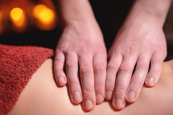 Close-up low key op vrouwen dij massage in een professionele premium spa salon, man masseur doet in een donkere kamer op de achtergrond van brandende kaarsen - Foto, afbeelding