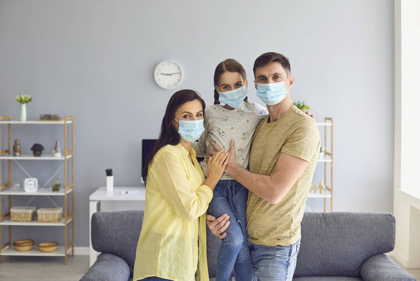 Family in medical masks on the face looks at the camera while standing in the room at home. - Fotoğraf, Görsel
