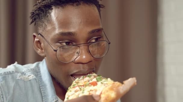 Portrait of an african young man eating a slice of pizza and drinking beer during the break - Imágenes, Vídeo