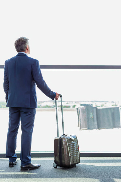 Portrait of mature businessman standing with his suitcase while looking through the window in airport - Foto, imagen