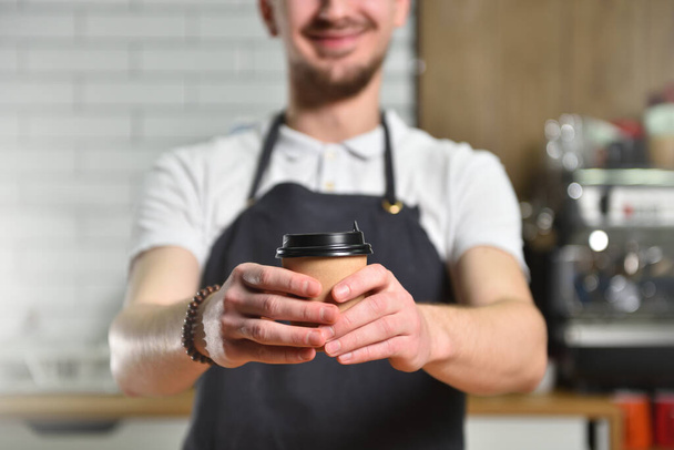 Young guy bartender barista holds out his hand with a paper cup of takeaway coffee. Text space - Photo, Image