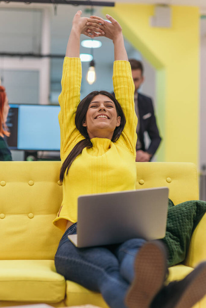 Przystojna bizneswoman (pracownik) siedzi na żółtej kanapie podczas pracy na laptopie w firmie startującej. - Zdjęcie, obraz