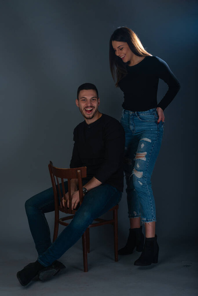 Handsome man sitting on a chair and beautiful woman standing next to him on dark background in a studio - Foto, Imagem