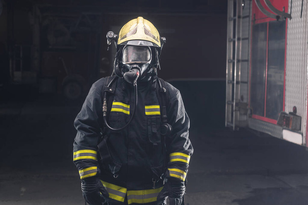 Portrait of a brave firefighter standing confident wearing full protective equipment, turnouts and helmet. Dark background with smoke and blue light. - Fotó, kép