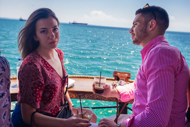 Beautiful couple sitting on the front of a boat on a calm blue sea and looking at the camera - Фото, изображение