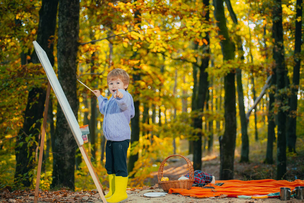 Pequeño niño pintando con caballete de dibujo en otoño otoño bosque parque fondo y sonriendo. Niño señalando con el dedo a la cámara. Gracioso niño feliz. Concepto de creatividad infantil
. - Foto, Imagen