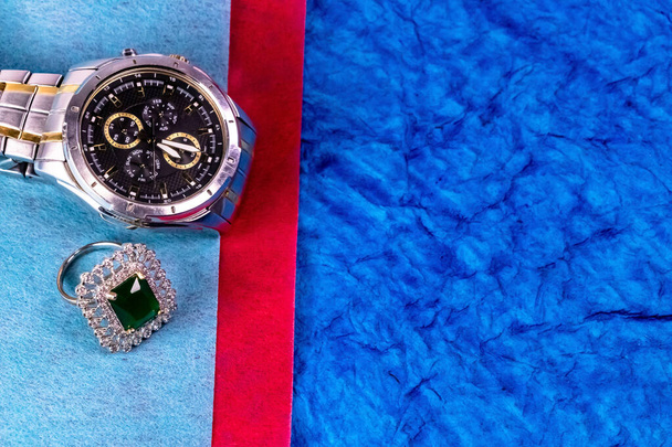 Primer plano de un reloj de pulsera de plata con anillo de esmeralda colocado en la superficie azul y rojo. concepto de aniversario de boda
 - Foto, Imagen