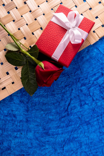 Portrait shot of a red gift box with one rose over blue and brown background. Wedding gifts concept - Valokuva, kuva