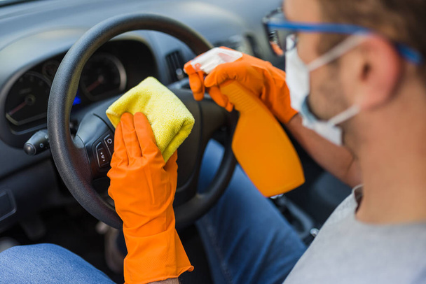 Worker with face mask cleaning car steering wheel with microfiber cloth and holding spray bottle of disinfectant. - 写真・画像