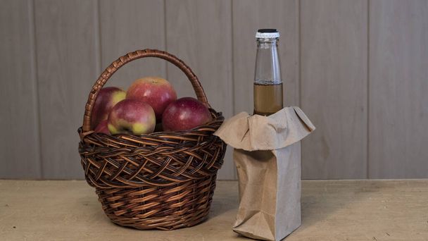 Basket with red fresh apples and a bottle of cider on a wooden table. - Photo, Image