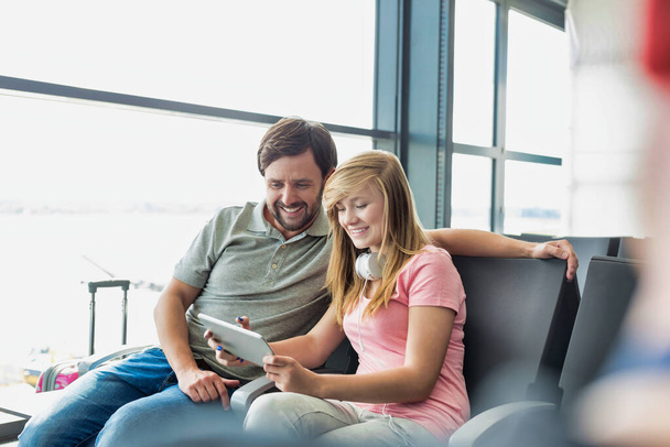 Portrait of young beautiful teenage girl showing digital tablet to her father while sitting and waiting for their flight in airport - Foto, Imagem