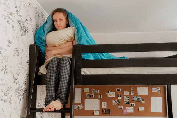 The guy is sitting on a bunk bed under a blue blanket and hugs a pillow, surprised, sad - Fotoğraf, Görsel