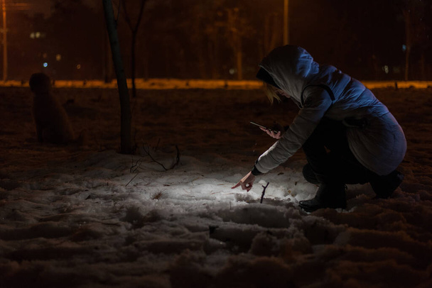 Μια γυναίκα ψάχνει για ένα χαμένο πράγμα από το φως ενός φακού έξω το χειμώνα. - Φωτογραφία, εικόνα