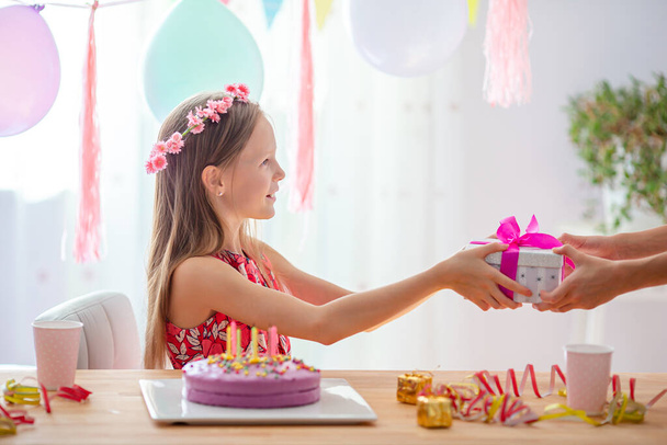 白人の女の子は夢のように笑って誕生日の虹のケーキを見ています。風船で祭りのカラフルな背景。誕生会と願いのコンセプト. - 写真・画像
