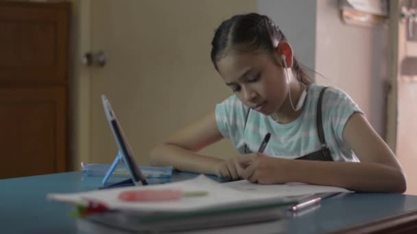 Adorable asian girl wearing earphones and using digital tablet for lesson online at home. Female teenager studying from home with video call during COVID-19 pandemic situation. - Imágenes, Vídeo