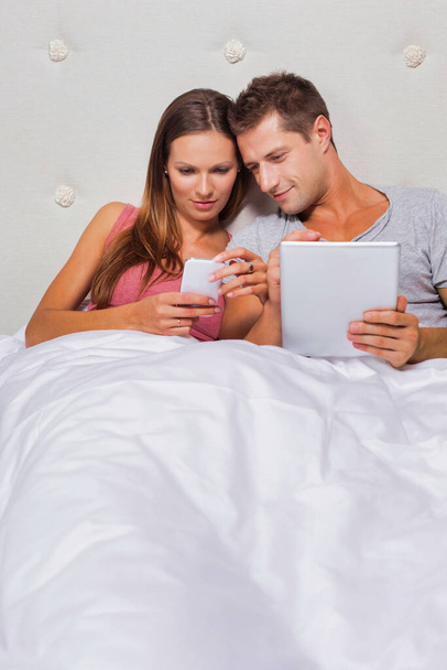 Portrait of young attractive woman showing smartphone while his boyfriend playing games on his digital tablet in bed - Valokuva, kuva