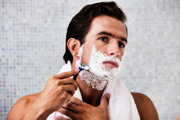 Portrait of young attractive man applying beard foam and shaving his beard while looking in the mirror - Foto, Imagem
