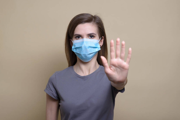 Porträt einer jungen Europäerin, die Maske trägt und Maske anbietet / gibt, während sie in die Kamera schaut. Beiger Hintergrund. Symptome einer Coronavirus-Erkrankung. Neuartiges Coronavirus (COVID-19)) - Foto, Bild
