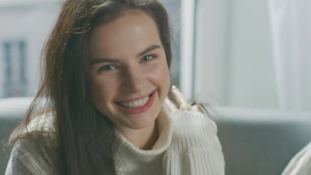 Portrait of Beautiful Young Brunette Smiling Charmingly while Looking at Camera, Brushing Her Lush Hair Away with a Flirtatious Gesture. Shy Girl Wearing White Knitted Sweater Laughing in Cozy Room - Filmati, video