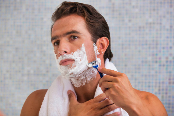 Portrait of young attractive man shaving his beard while looking in the mirror - Foto, Imagem