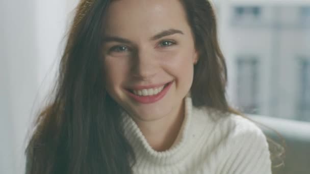 Portrait of Brunette Smiling Charmingly while Sitting on Sofa, Brushes Her Lush Hair Away with Flirtatious Gesture. Girl Wearing Oversized White Knitted Sweater Relaxes on Couch in Her Cozy Apartment - Filmagem, Vídeo