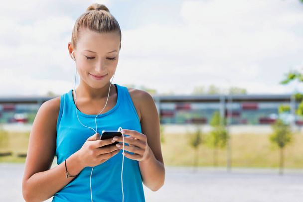Portrait of young attractive woman standing while using and listening to music on her smartphone in park - Photo, Image