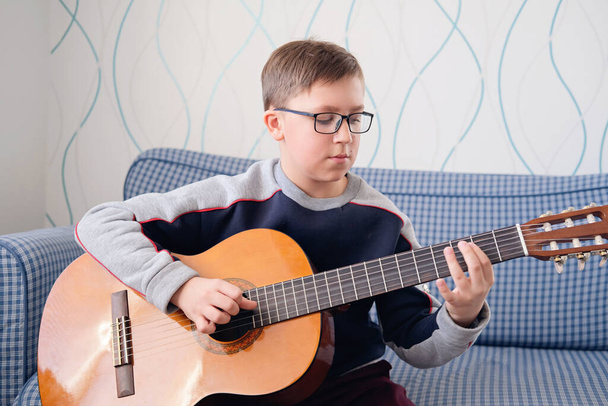 ギターを弾くことを学ぶ。音楽教育。アコースティックギターを弾く少年。屋内でプレーしようとしているメガネの深刻なティーンエイジャー. - 写真・画像