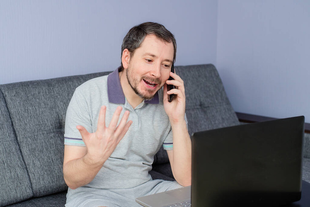 A man works at home behind a laptop and talks on the phone - Photo, Image