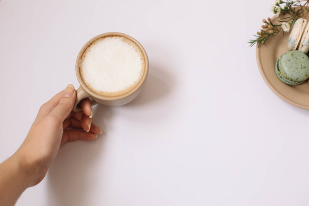 Macarons coloridos caseiros na placa marrom no fundo branco. Mão feminina está segurando um copo de café.Blossom
. - Foto, Imagem