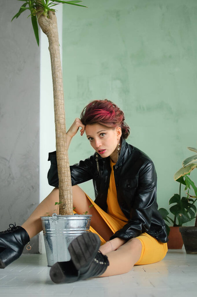 Beautiful girl in glam rock style with dark pink hair and mohawk in yellow dress and black leather jacket is sitting on the floor near a flower pot with houseplant - Photo, Image