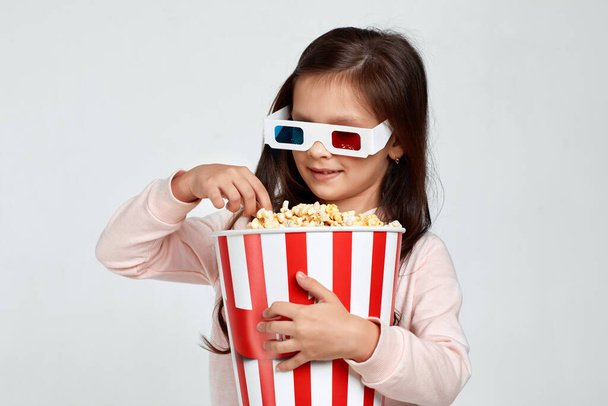 child girl in red-blue 3d glasses holding popcorn bucket - Фото, изображение
