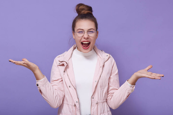 Image of charismatic adorable attractive lady with bun standing isolated over lilac background, opening mouth widely, raising arms, being in high spirits, wearing casual clothes. Emotions concept. - Fotoğraf, Görsel