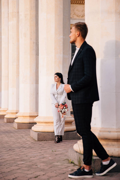bride and groom in a white pantsuit posing on the background of large white columns - Foto, afbeelding