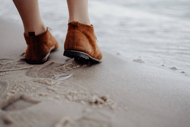 Os pés das mulheres em botas de couro marrom em uma praia de areia deixam pegadas na areia molhada em um dia nublado
. - Foto, Imagem