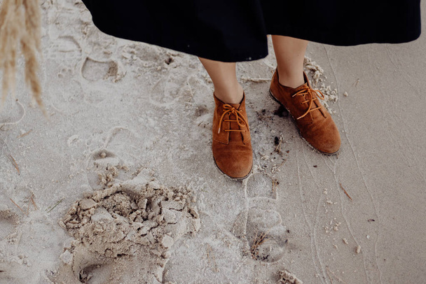 A girl in brown leather boots stands on a wet sandy seashore on a cloudy day with dry decorative grass in her hands - Foto, Imagen
