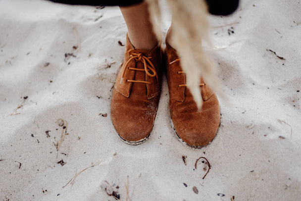 Uma menina em botas de couro marrom fica em uma praia de areia molhada em um dia nublado com grama decorativa seca em suas mãos
 - Foto, Imagem