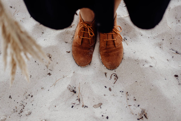 Uma menina em botas de couro marrom fica em uma praia de areia molhada em um dia nublado com grama decorativa seca em suas mãos
 - Foto, Imagem