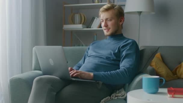 Portrait of Handsome Blonde Young Man Working on a Laptop Computer, While Sitting on a Chair in His Cozy Living Room. Creative Freelancer Relaxes at Home, Surfs Internet, Uses Social Media and Relaxes - Video