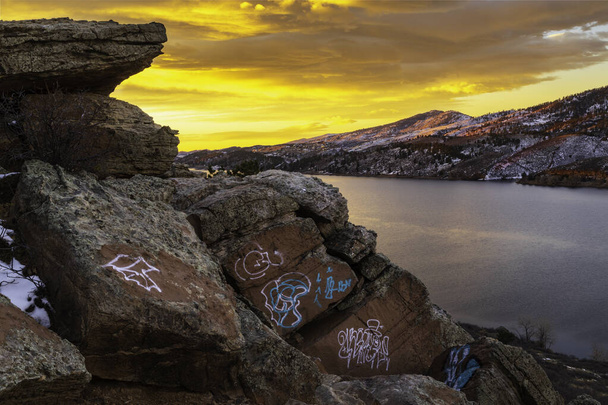 The sky bursts with color on Horsetooth Reservoir with graffiti on the boulders overlooking the lake - Photo, Image